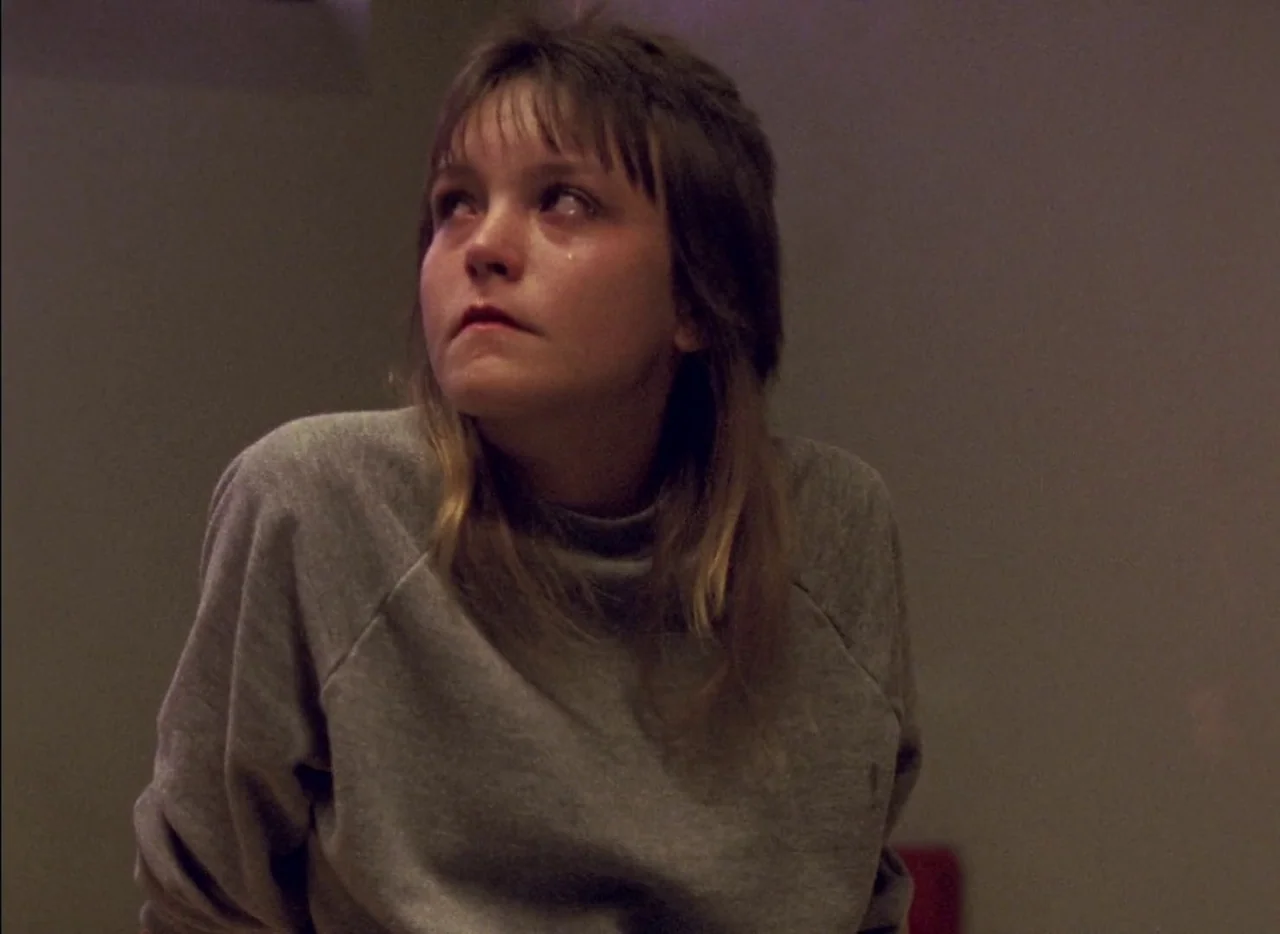 Streetwise (1984) movie still: medium — A dimly lit room with an indistinct neutral-toned wall in the background. A young woman with damp eyes looks…