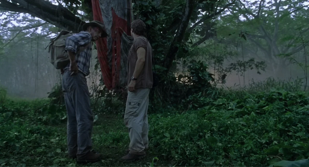 Jurassic Park III (2001) movie still: fullbody — A dense, foggy tropical environment filled with overgrown plants and large trees. Two men stand in…