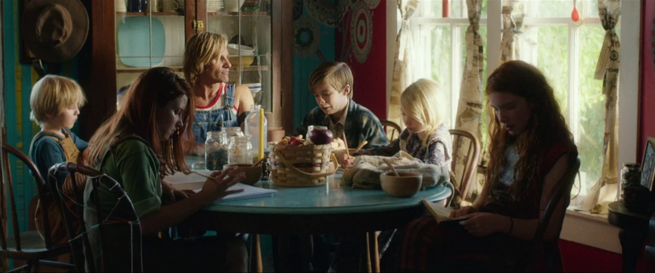 Captain Fantastic (2016) movie still: wide — A rustic room features red walls with circular art and a large window. A family sits around a round…
