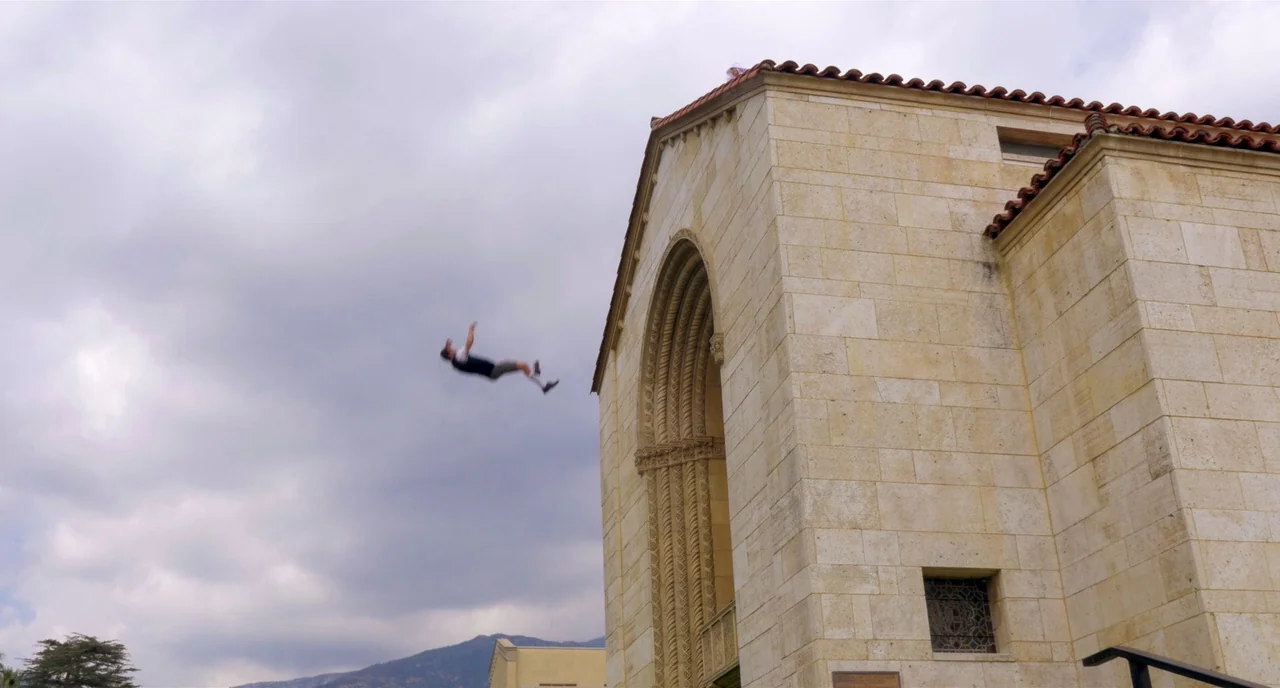 Nightmare Cinema movie still: wide — A stone building with a large Romanesque arch stands against a cloudy sky. A person is airborne, falling…