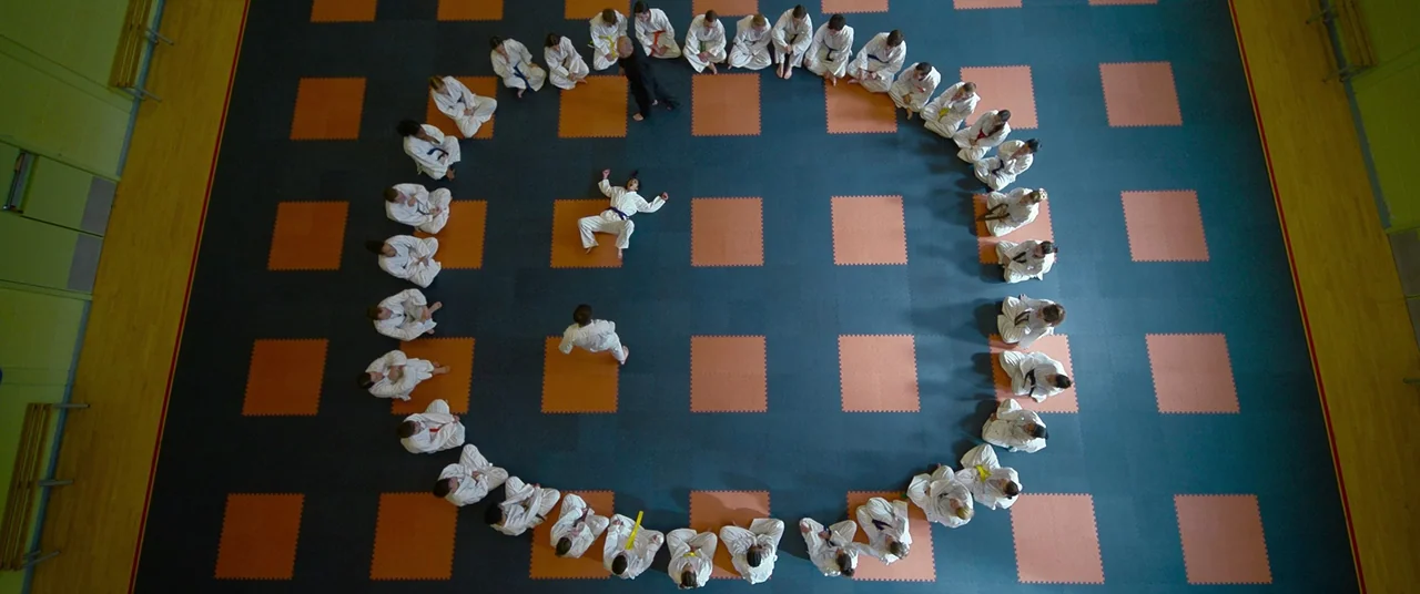 Polite Society (2023) movie still: wide — A top-down view shows a gymnasium floor covered with a grid of dark teal and orange square mats. A large…