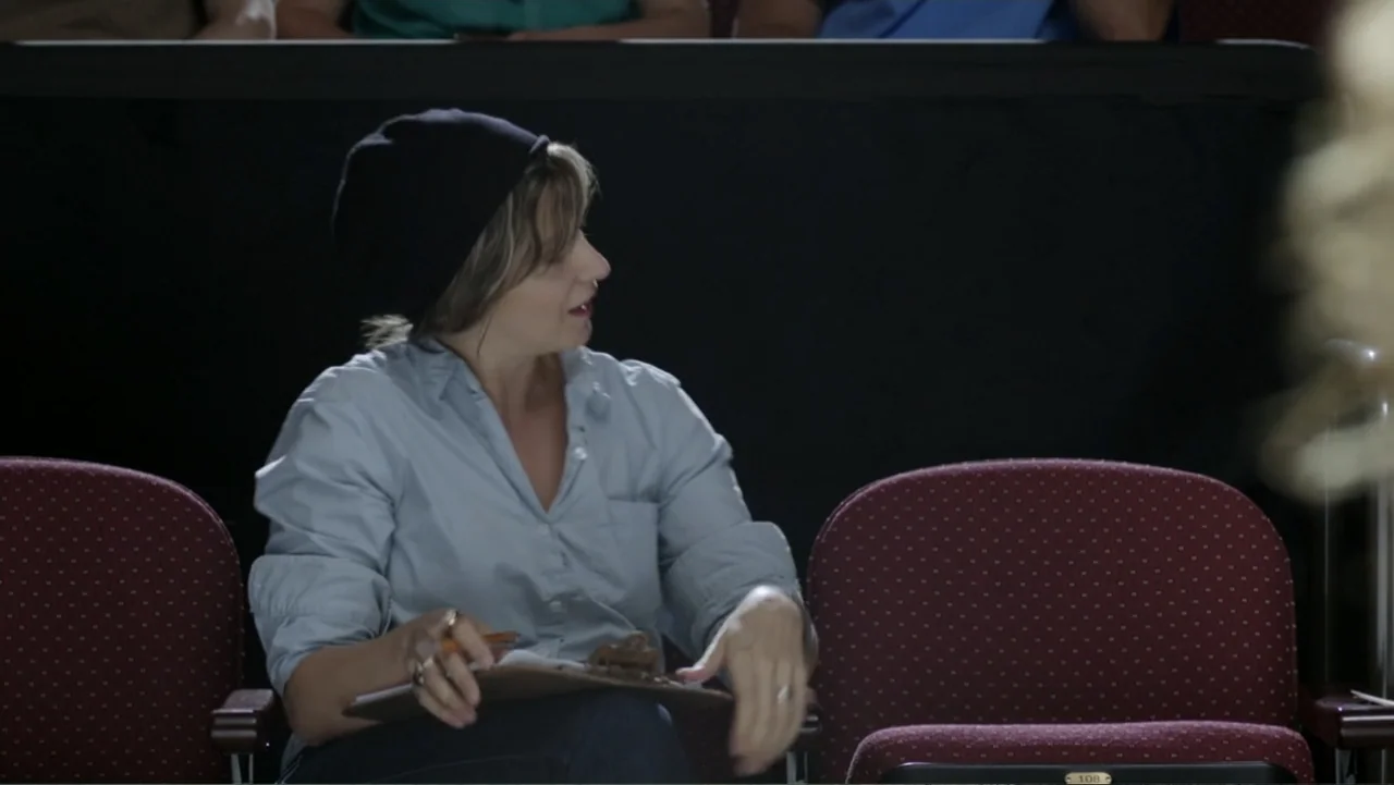 At Middleton (2013) movie still: medium — An auditorium with rows of padded dark red seats arranged in a dark space. A woman sits in the center seat…