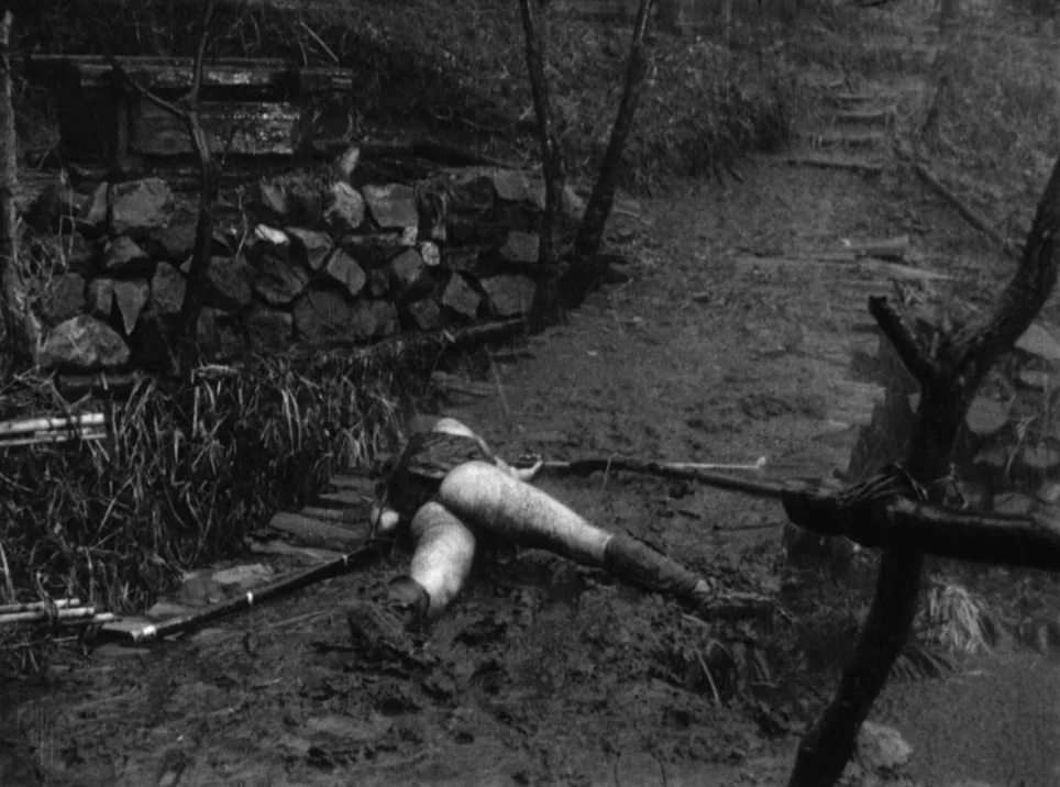Seven Samurai (1954) movie still: wide — A rain-soaked muddy trail in a wooded area borders a stacked stone retaining wall. A male body lies face…
