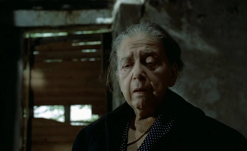 Lacombe, Lucien (1974) movie still: closeup — An elderly woman stands before a dark, textured wall in a worn interior space. Behind her, a partially…