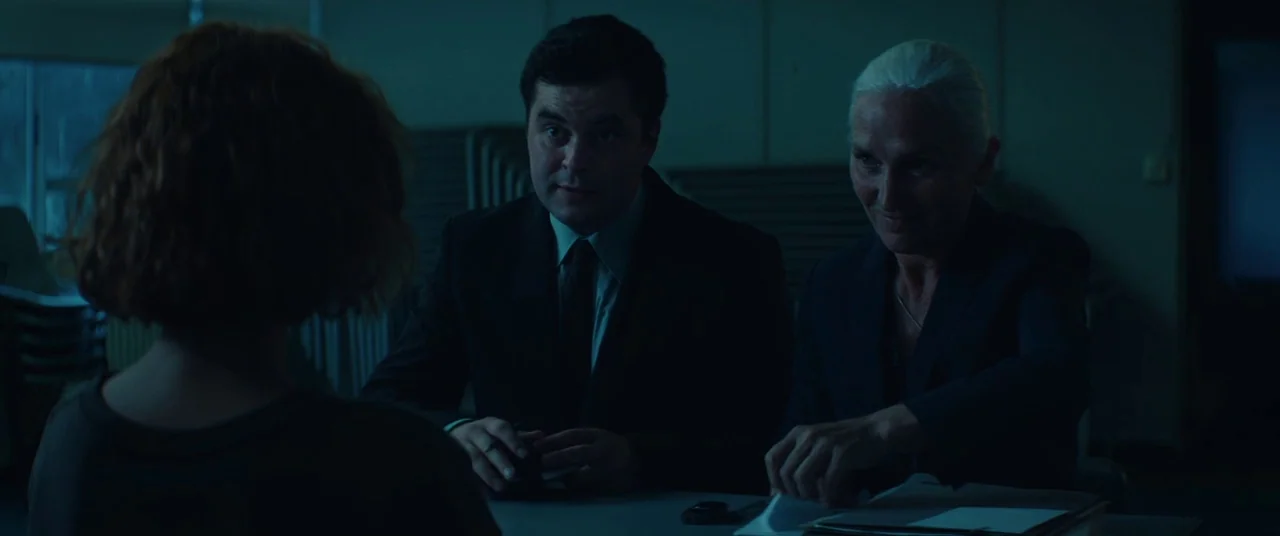 Beast (2017) movie still: medium — A dark, dimly lit room contains two investigators sitting at a table. The two individuals face a person whose…