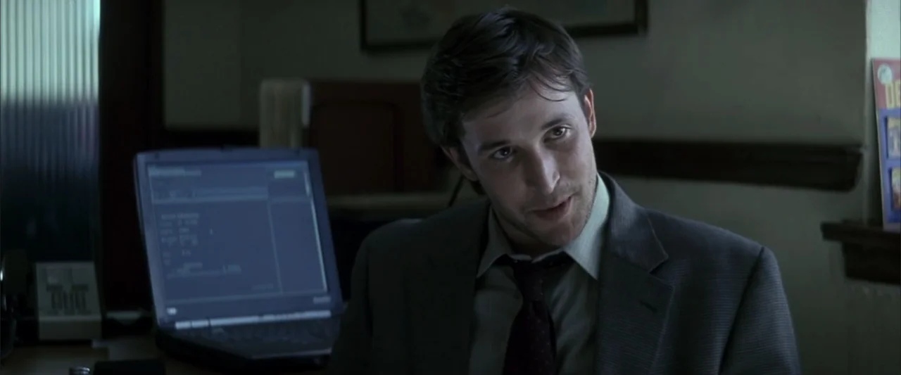 Enough (2002) movie still: medium — A dark office environment features a desk with a laptop glowing in the foreground. A man in a suit sits at the…
