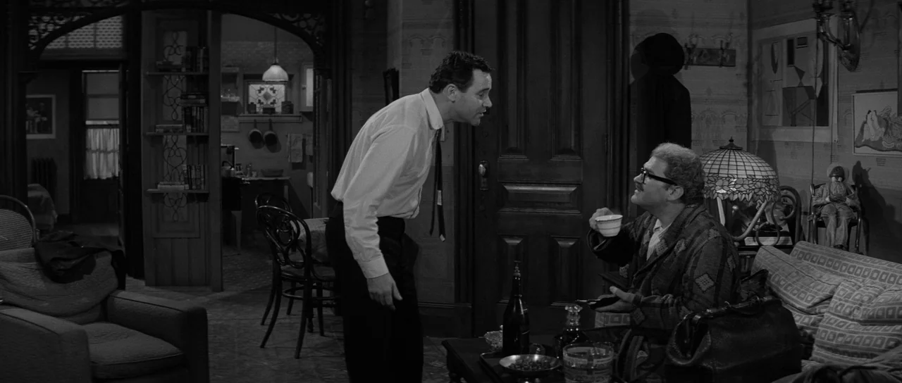 The Apartment (1960) movie still: medium — A dark living room with vintage furniture and a large wooden doorway. One man stands while speaking to a…