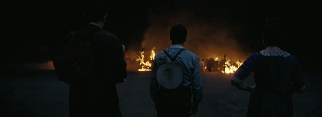 Sinners (2025) movie still: fullbody — An open dark field at night is illuminated by several large distant bonfires. Three people stand in a line…