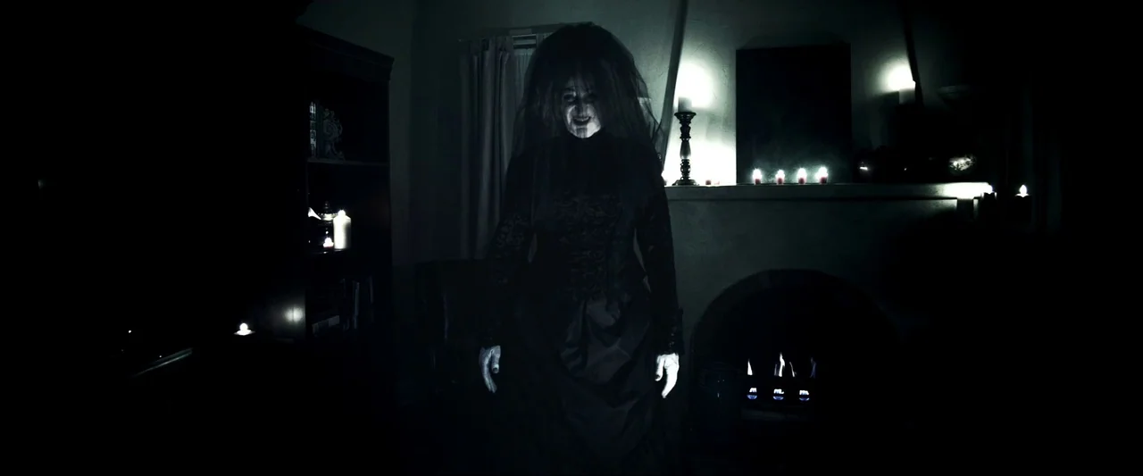 Insidious (2010) movie still: fullbody — A dark living room features a fireplace with flickering flames and numerous lit candles. A woman in a black…