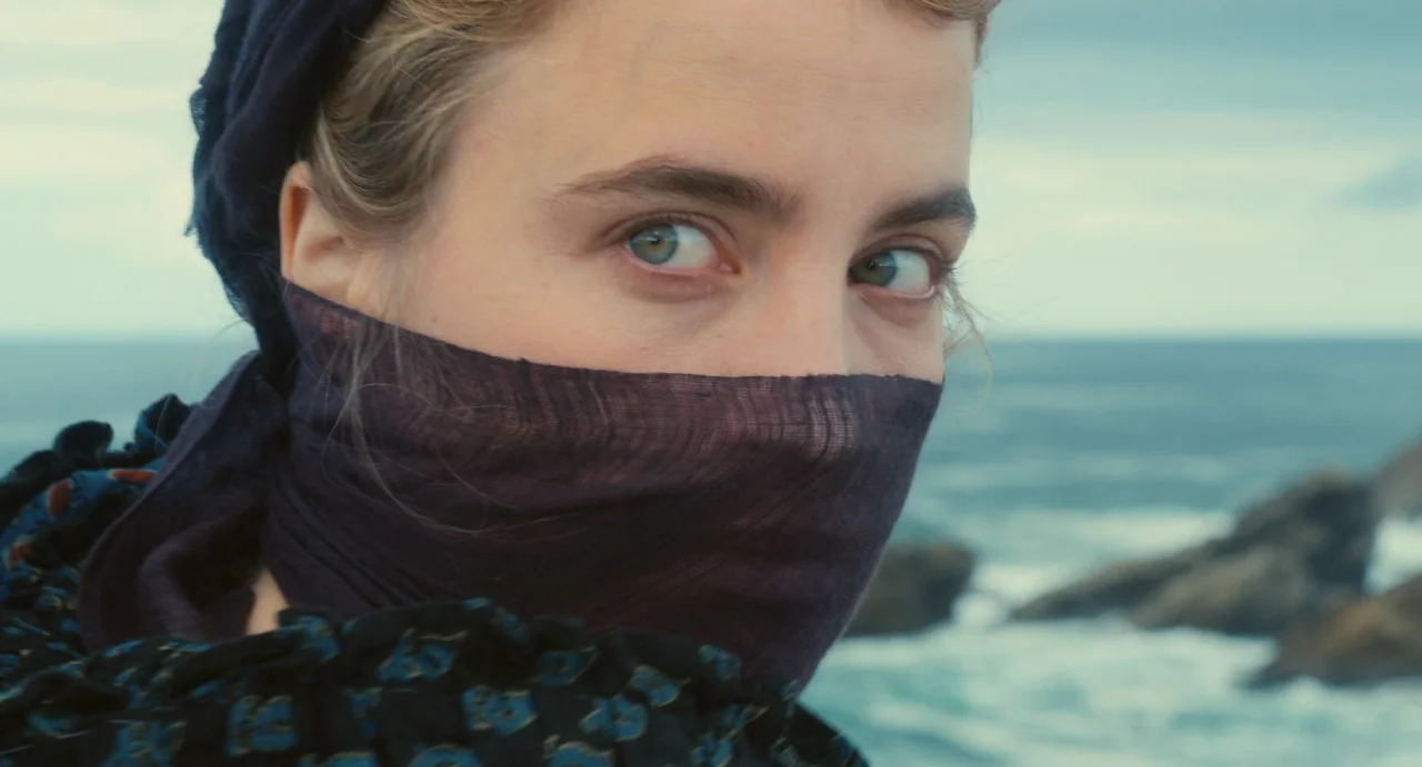 Portrait of a Lady on Fire movie still: closeup — A woman stands near a rocky coastline with ocean waves visible in the background. She wears a dark…