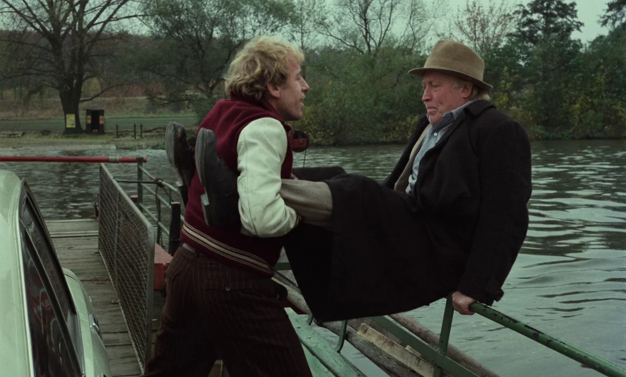 Wrong Move (1975) movie still: fullbody — A wooden ferry deck with a metal railing sits on calm, murky water. A younger man in a letterman jacket…