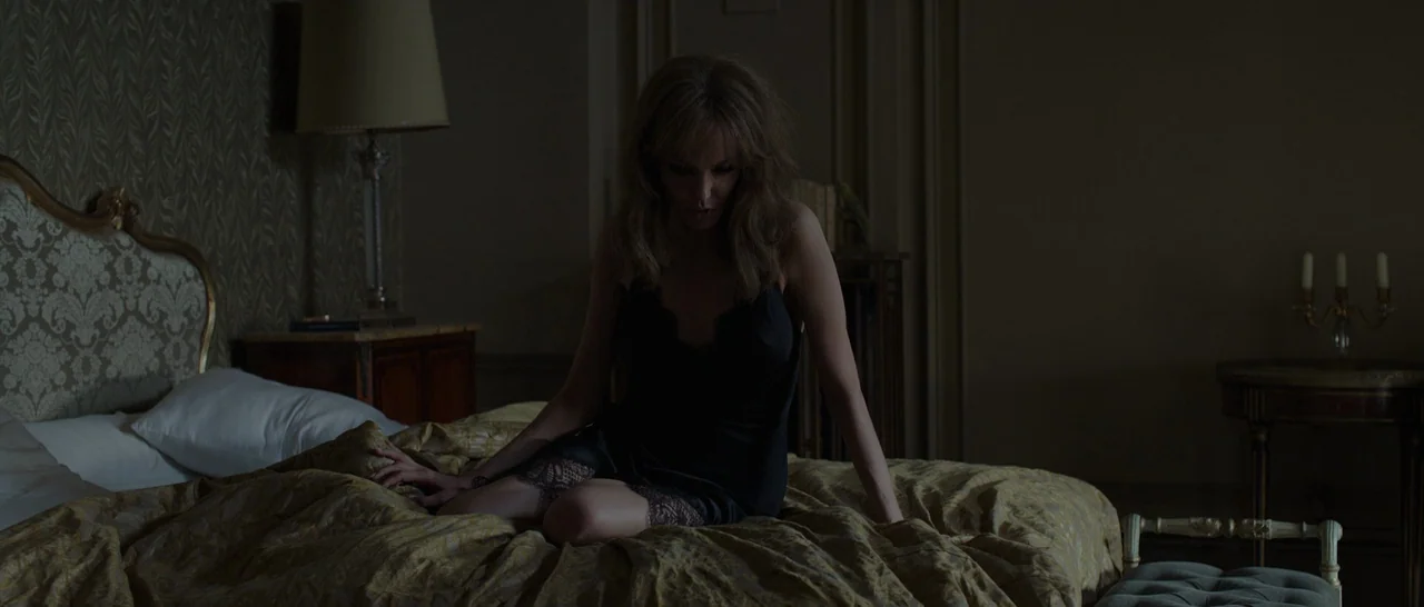 By The Sea (2015) movie still: medium — A dark hotel room features vintage patterned wallpaper and ornate, classic furniture pieces. A woman sits on…