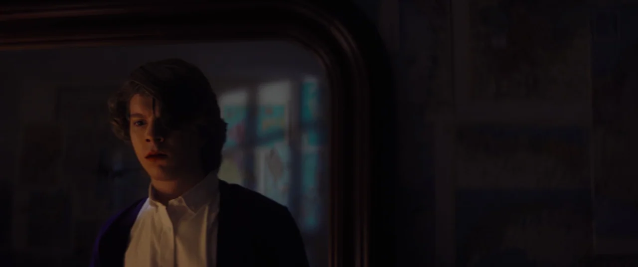 Linoleum (2022) movie still: medium — A dark indoor space contains a large wooden framed mirror hanging on the wall. A young man stands in front of…