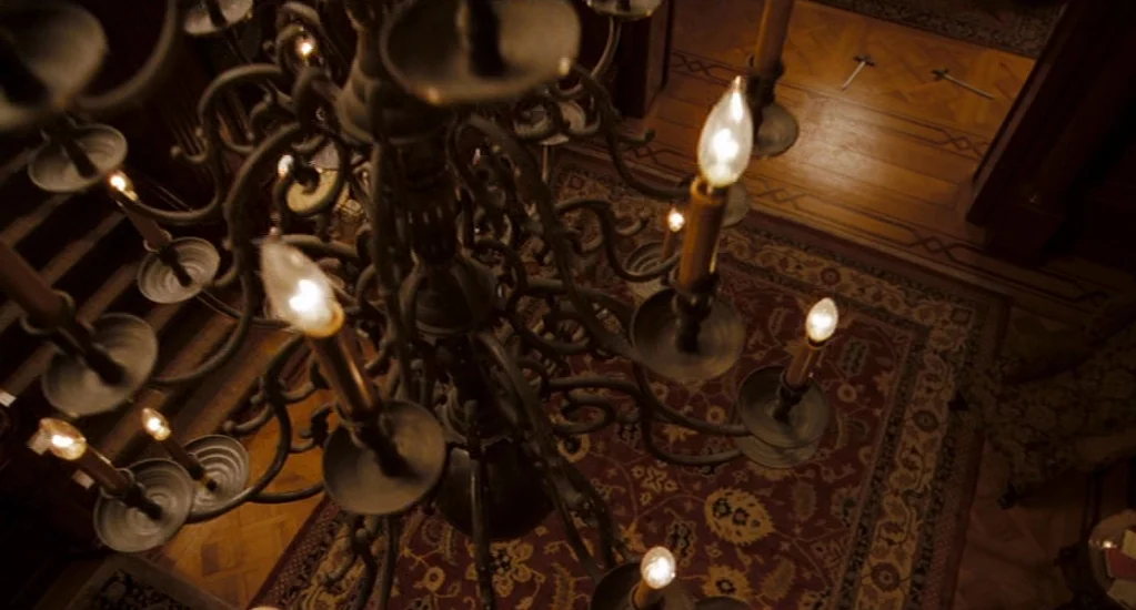 Valkyrie (2008) movie still: wide — An ornate dark iron chandelier hangs over a patterned rug in a wood-floored hall. Two narrow swords lie crossed…