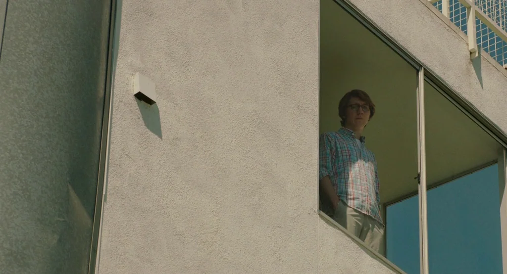 Ruby Sparks (2012) movie still: medium — A modern building exterior features textured concrete walls and large glass window sections. A man stands…