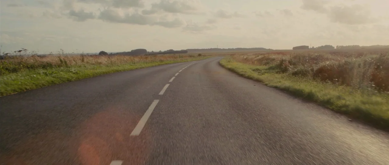 Road Games (2015) movie still: establishing — A quiet asphalt road curves through flat, grassy fields under a cloudy sky. The horizon is low and the…