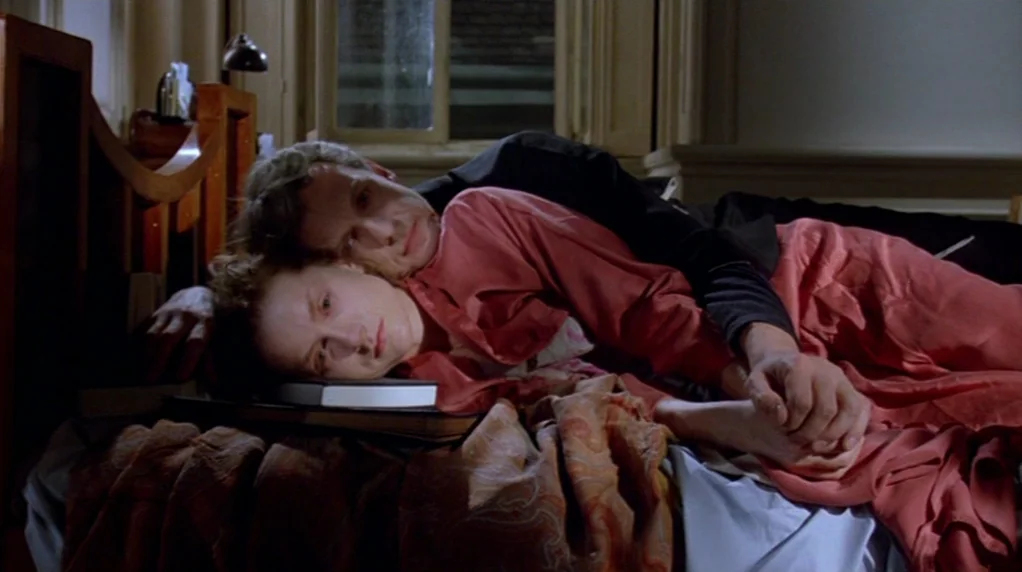 Malina (1991) movie still: medium — A dimly lit room with wooden furniture and a window showing dark night outside. A man and woman rest…