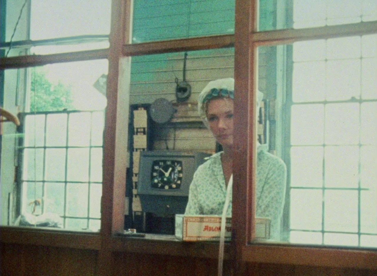 Wanda (1970) movie still: medium — A woman wearing a head covering sits behind a glass partition in a workspace. A vintage wall clock and a small…