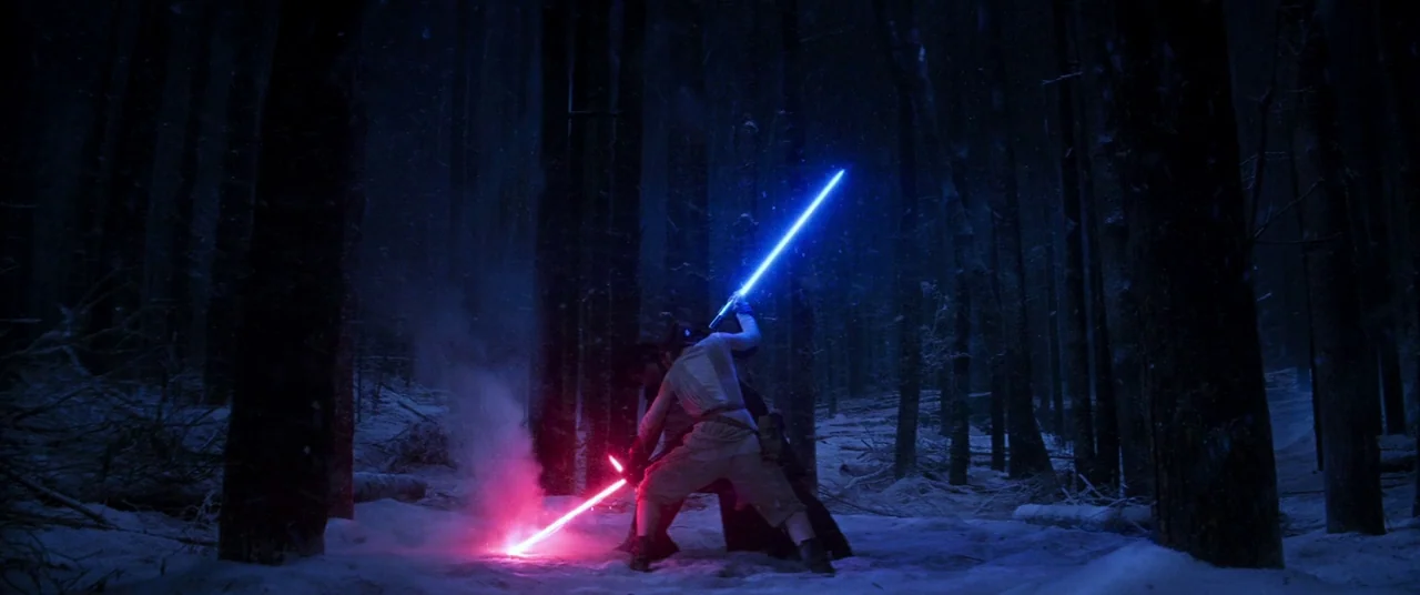 Star Wars: The Force Awakens (2015) movie still: wide — A dark, snow-covered forest contains tall, bare trees at night. Two combatants clash sabers,…