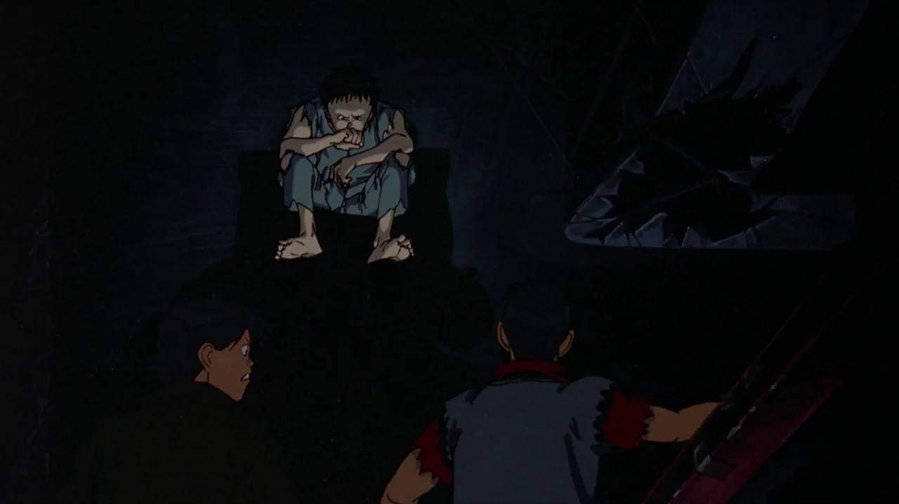 Akira (1988) movie still: medium — A dark, industrial underground space with uneven surfaces and structural rubble. Two youths approach a lone…