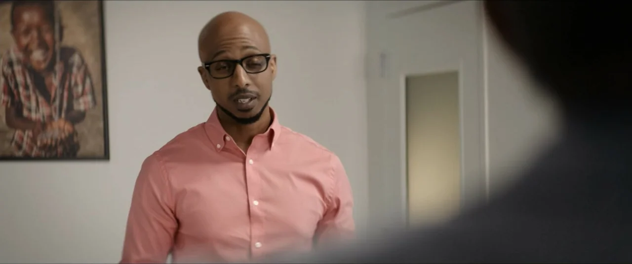 The Forge (2024) movie still: medium — A brightly lit modern office interior with a photograph of a child hanging nearby. A bald man wearing glasses…
