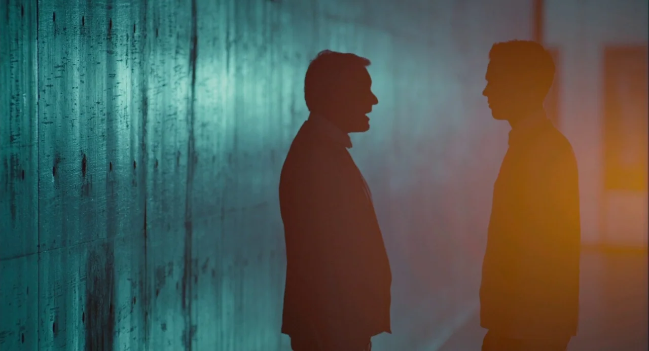 Equals (2015) movie still: medium — A narrow corridor features a long board-marked concrete wall on the left side. Two men stand in silhouette…