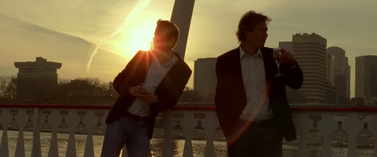 Mississippi Grind (2015) movie still: medium — Two men stand at a metal railing overlooking a river at sunset. Buildings are visible on the horizon…