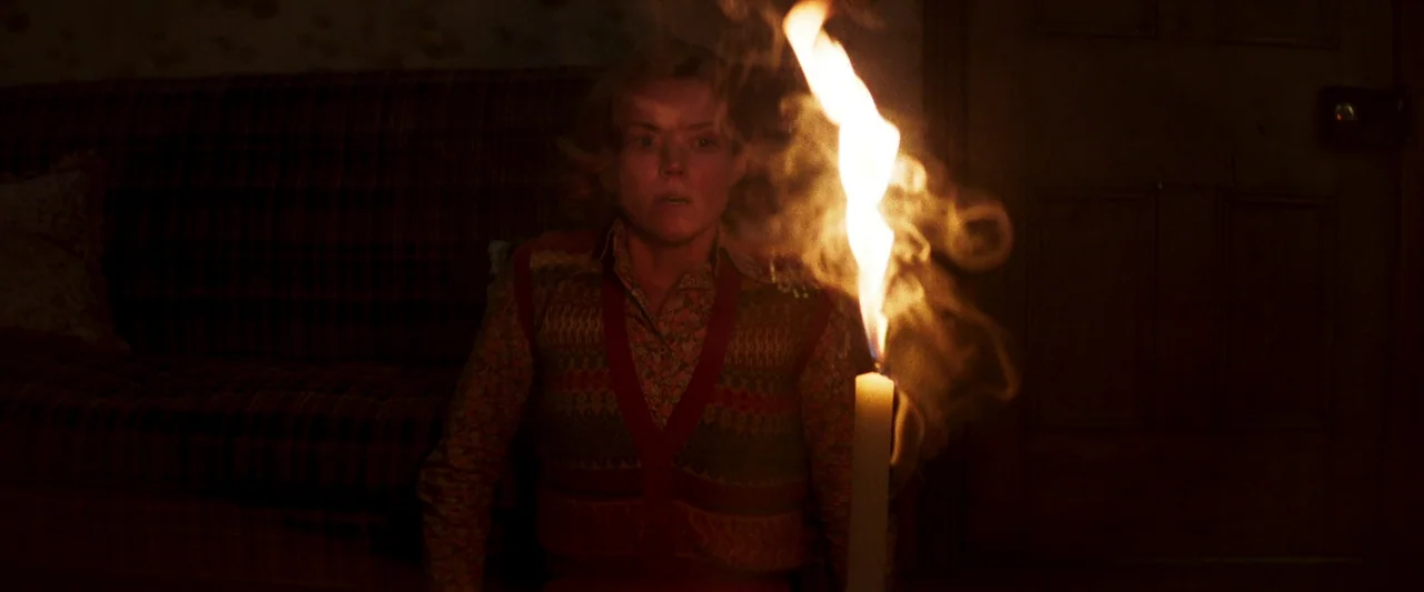 Starve Acre (2023) movie still: medium — A dim, dark room features patterned wallpaper and a closed wooden door. A blonde woman sits in front of a…