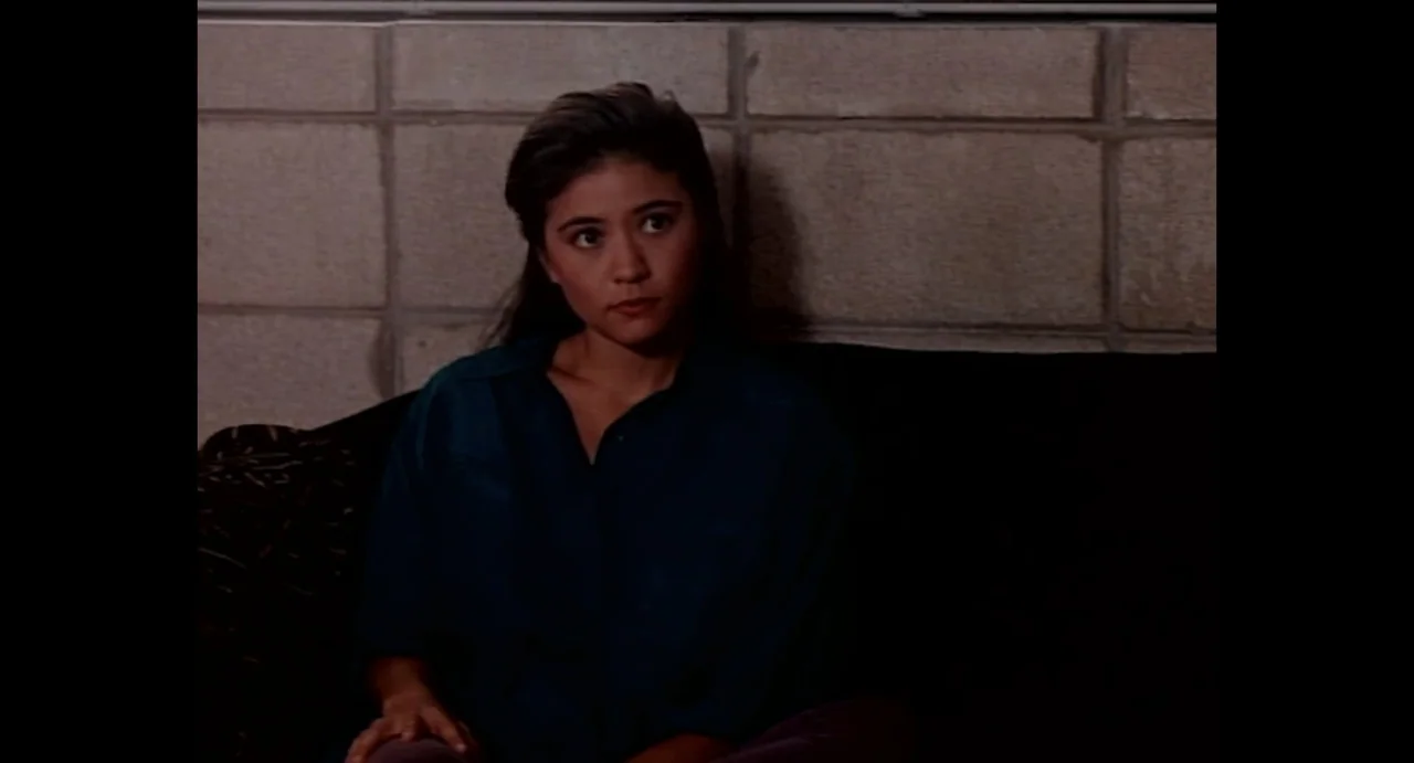 American Yakuza (1993) movie still: medium — A woman sits on a sofa in front of a concrete block wall. She looks toward the side while resting on…