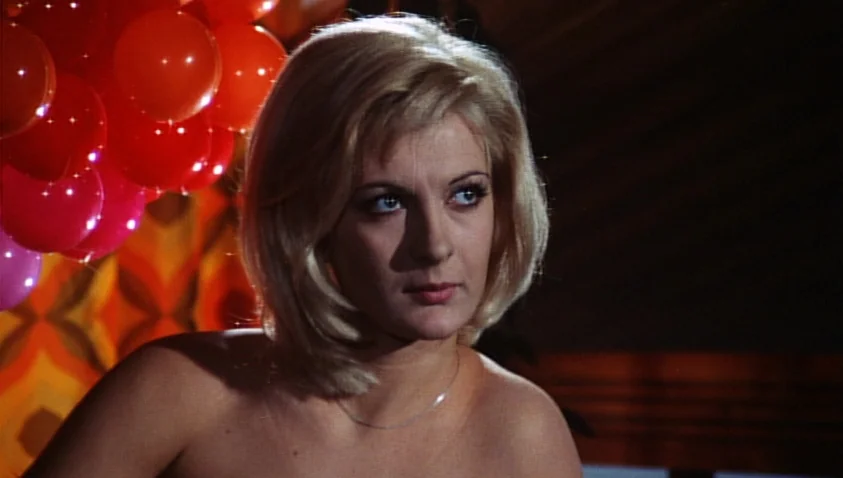 A Bay of Blood (1971) movie still: closeup — A dimly lit room features a patterned wall and a cluster of red balloons. A blonde woman with a bob…