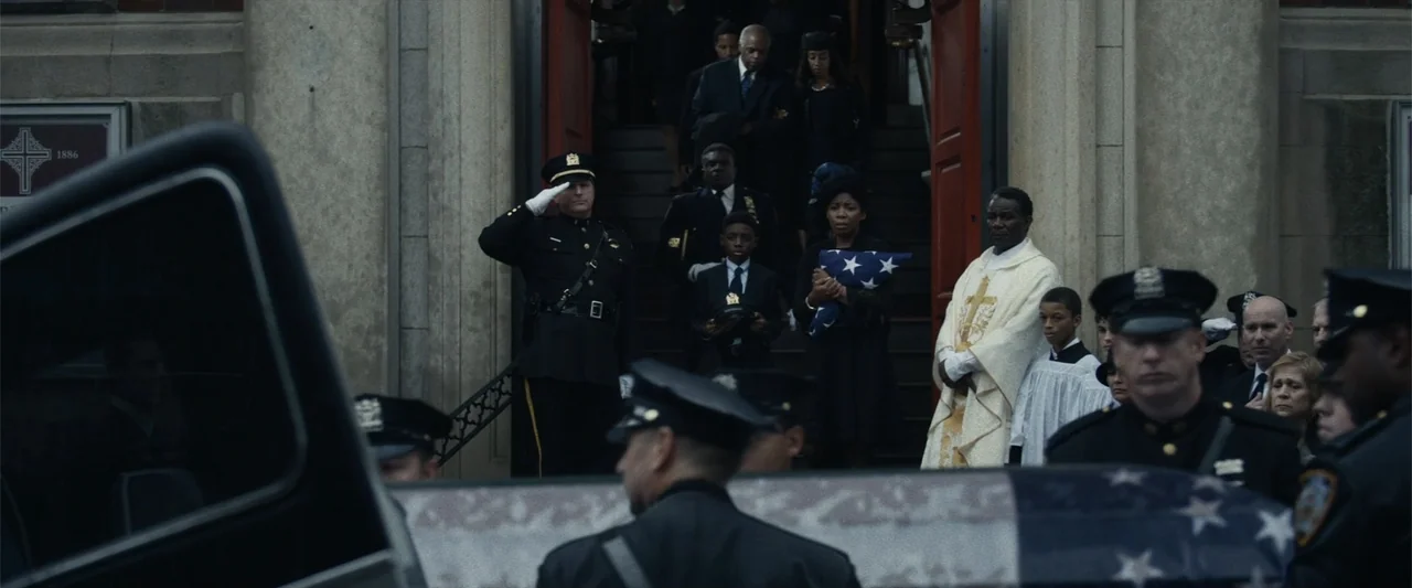 21 Bridges (2019) movie still: fullbody — A group of mourners exits a stone church building down a flight of stairs. Police officers stand in…
