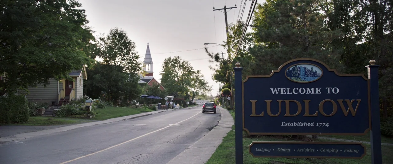 Pet Sematary (2019) movie still: establishing — A quiet small town street features a welcome sign in the immediate foreground. In the background a…