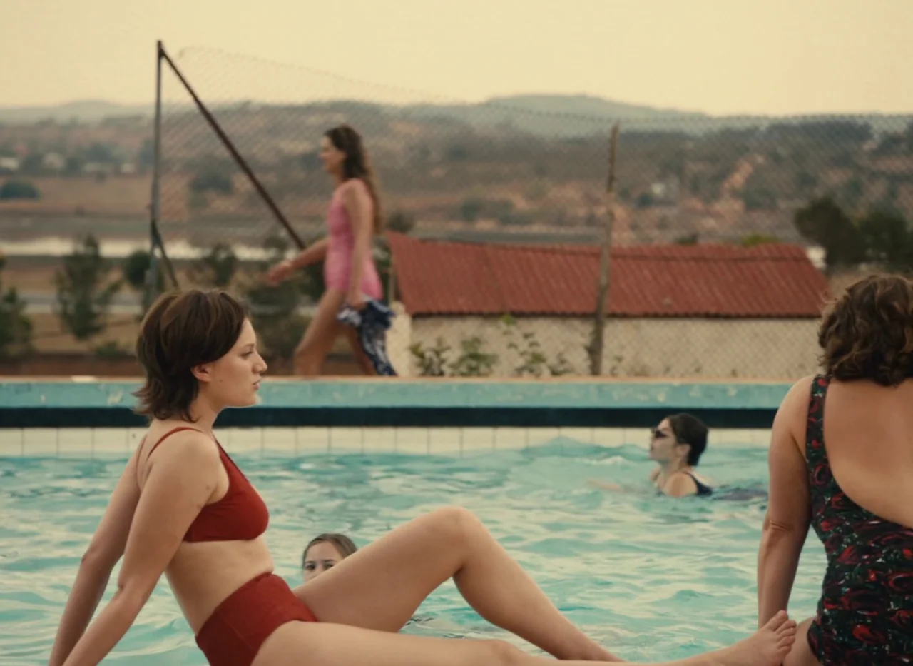 Red Island (2023) movie still: medium — A public outdoor pool area features a concrete deck and a chain link fence. Several people are relaxing in…