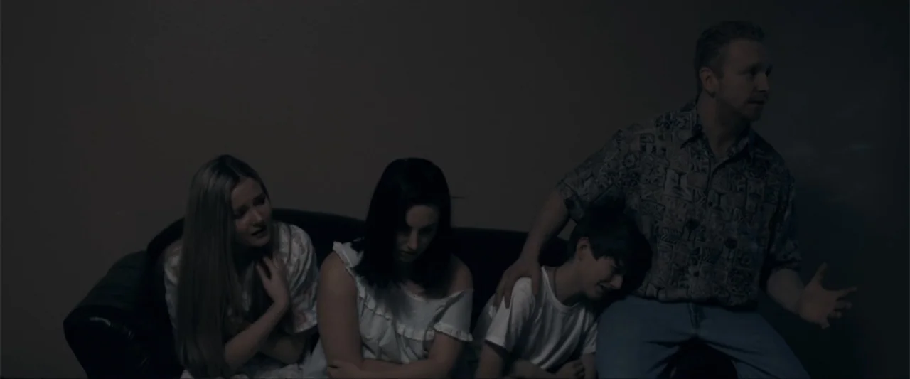 Anomaly (2006) movie still: medium — A dimly lit room features four people sitting on a dark couch together. A man rests his hand on a crying boy's…