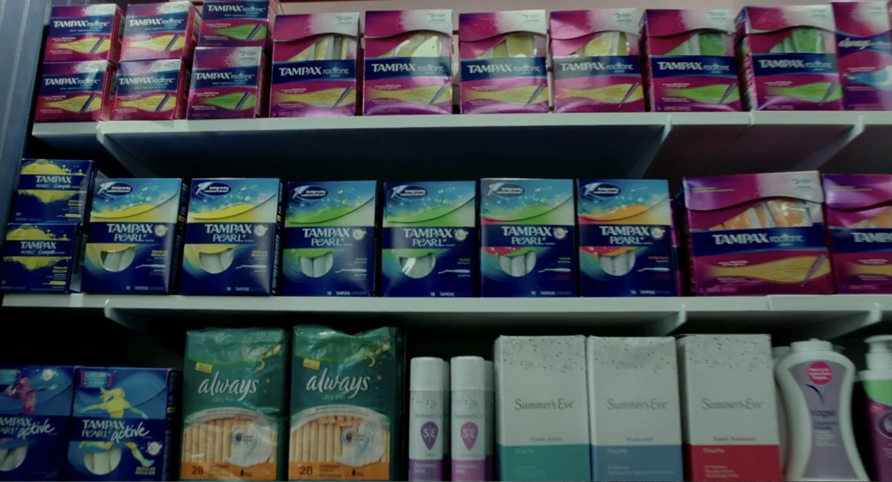 Blended (2014) movie still: medium — Shelves in a retail environment are stocked with various feminine hygiene product boxes. The items are arranged…