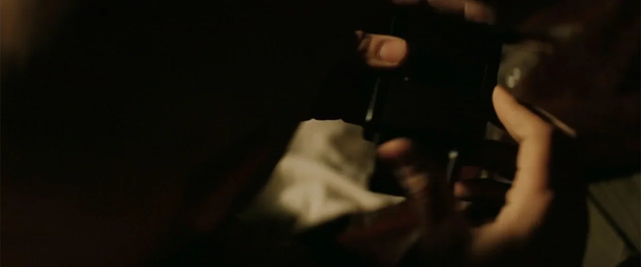 xXx (2002) movie still: closeup — A dimly lit interior room features dark shadows and soft textural highlights on skin. Two hands manipulate a small…