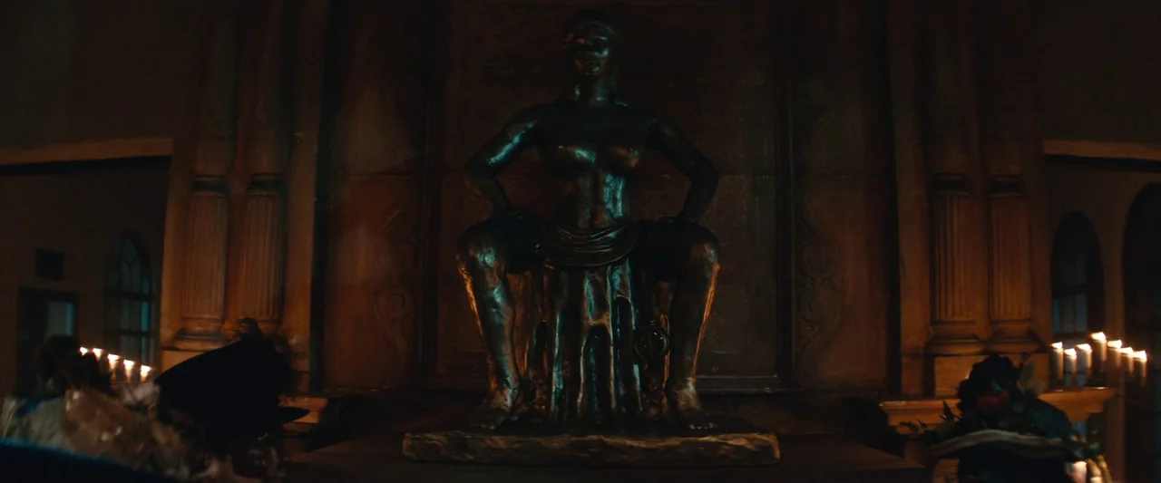 The Mad Hatter (2021) movie still: medium — A bronze sculpture sits on a pedestal in a dimly lit, shadowy room. The sculpture depicts a female…