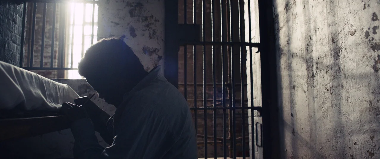 Judas and the Black Messiah (2021) movie still: medium — A dimly lit room features textured peeling paint on concrete walls and vertical iron bars.…