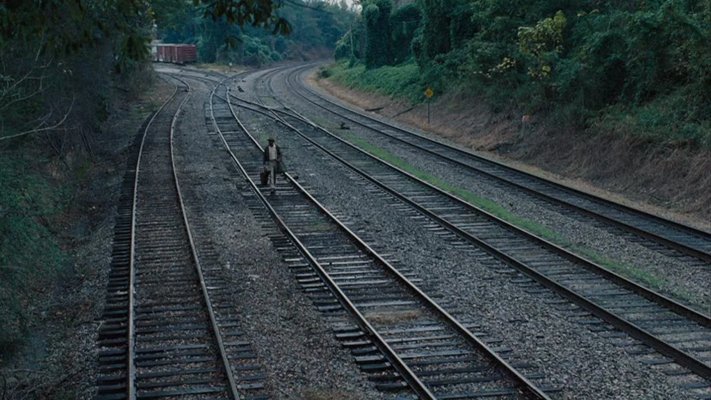 Honeydripper (2007) movie still: wide — A set of four train tracks runs through a wooded, rural area. A lone man walks along the center tracks…
