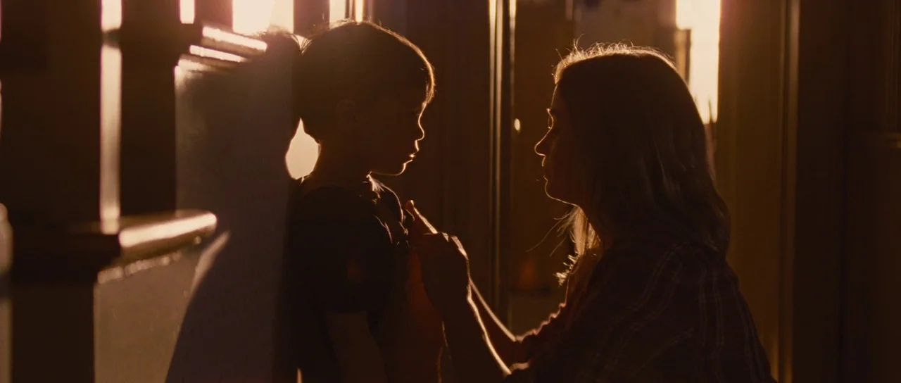 Looper (2012) movie still: medium — A residential hallway with dark walls illuminated by bright light from an adjacent window. A woman faces a small…