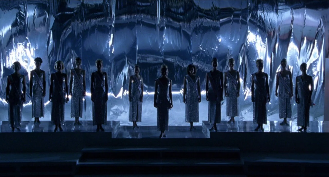 Drop Dead Gorgeous (1999) movie still: wide — A row of twelve women stands on a raised platform before a wrinkled silver curtain. The subjects are…