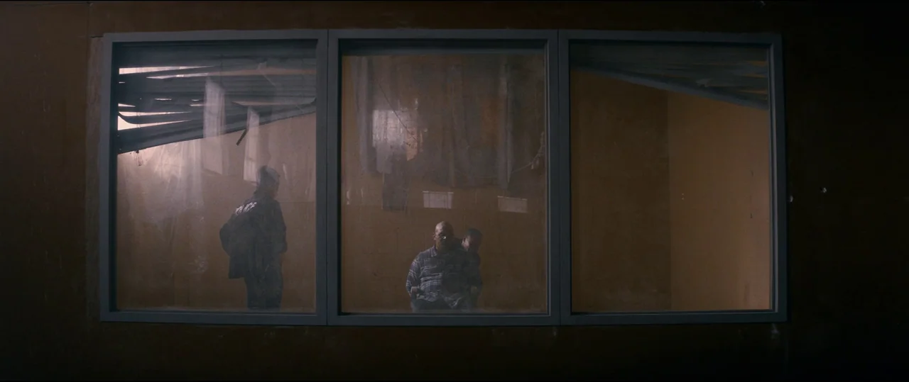 El Chicano (2018) movie still: wide — A dimly lit room viewed through a large three-pane window frame. One person stands and two others sit in the…