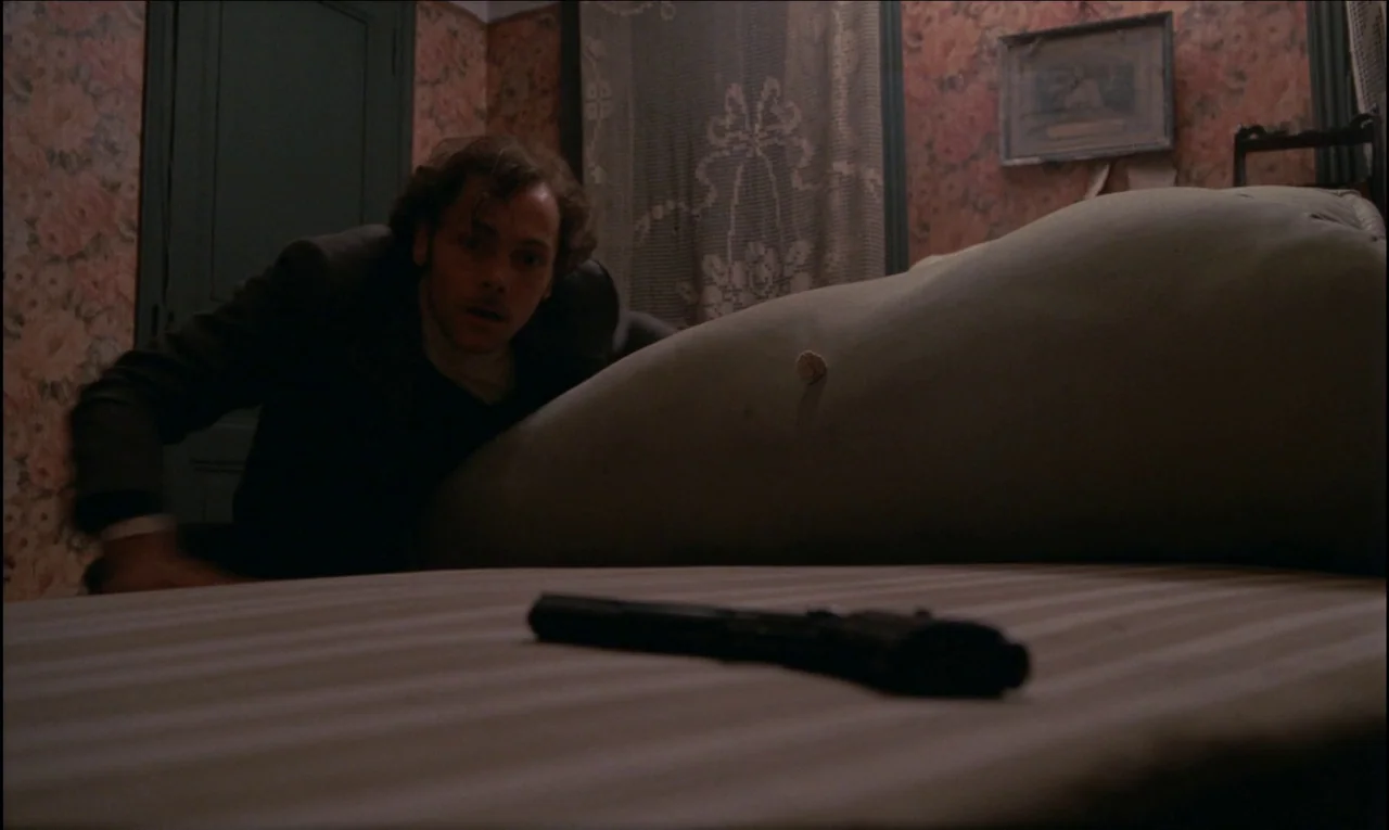 Serie Noire (1979) movie still: medium — A small room features floral wallpaper, lacy curtains, and a low-slung bed frame. A man hides behind the…