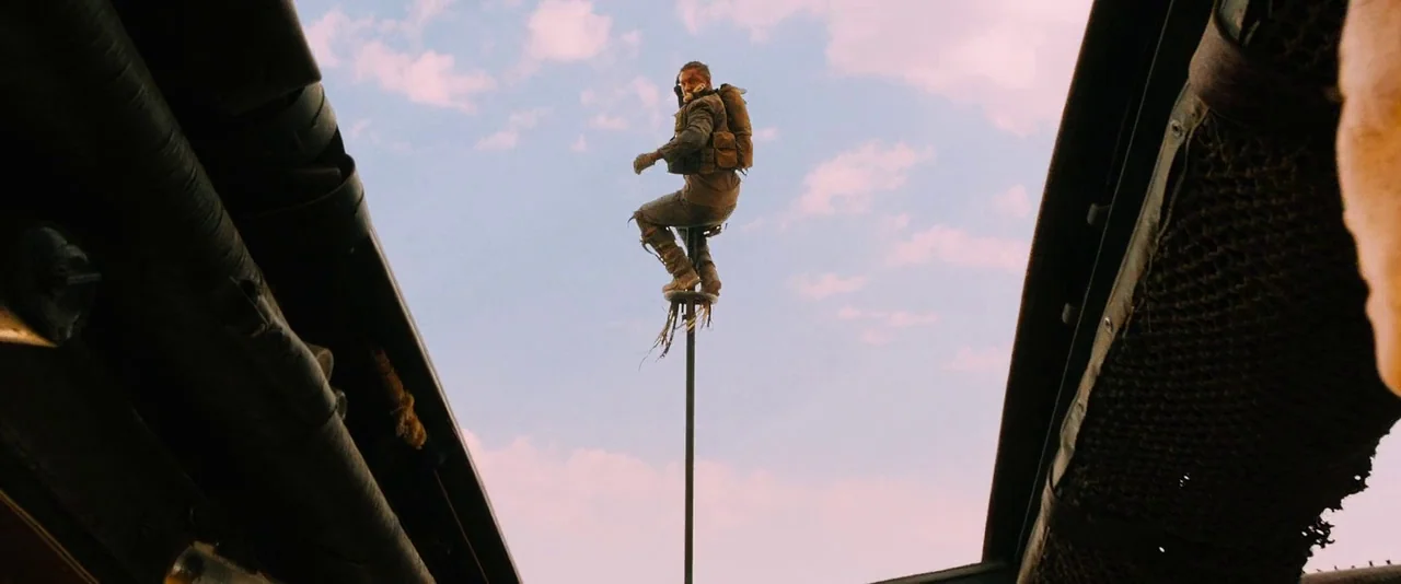 Mad Max: Fury Road (2015) movie still: wide — A man stands atop a tall, swaying metal pole extending from a moving vehicle. The background shows a…