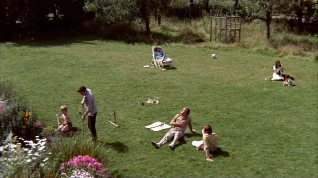 Accident (1967) movie still: wide — A sprawling green lawn contains a cluster of people engaged in leisurely afternoon activities. A man stands with…