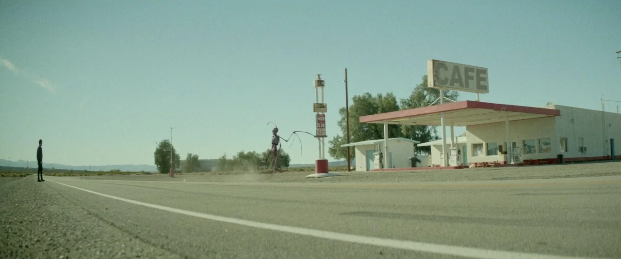 Southbound (2015) movie still: wide — A remote desert landscape features an abandoned gas station with a large cafe sign. A lone man stands on the…