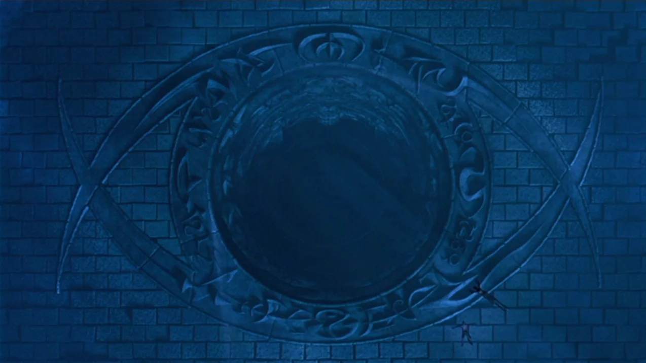 Dagon (2001) movie still: establishing — A massive eye-shaped stone structure is embedded in a vast brick floor. Two small human figures lie on the…