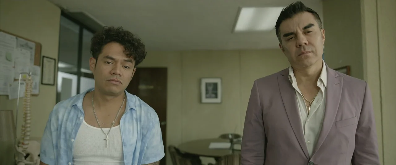Divine Intervention (2026) movie still: medium — A sterile office features light green walls and a small dark table behind them. Two men stand in…