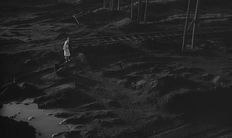 Man Is Not Bird (1965) movie still: wide — A dark, industrial railway yard features uneven mounds of dirt and exposed metal tracks. Two small…