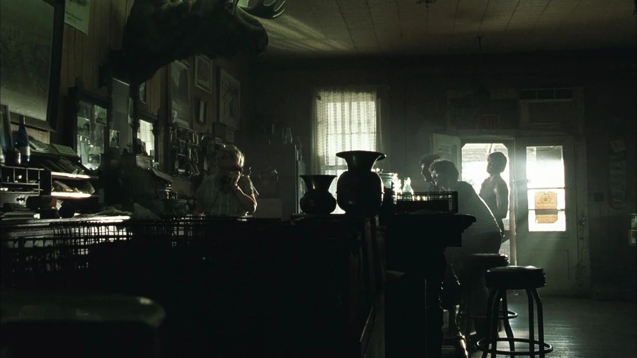 The Texas Chainsaw Massacre (2003) movie still: wide — A dimly lit rural store with wood paneling and a large moose head mount. An elderly woman…
